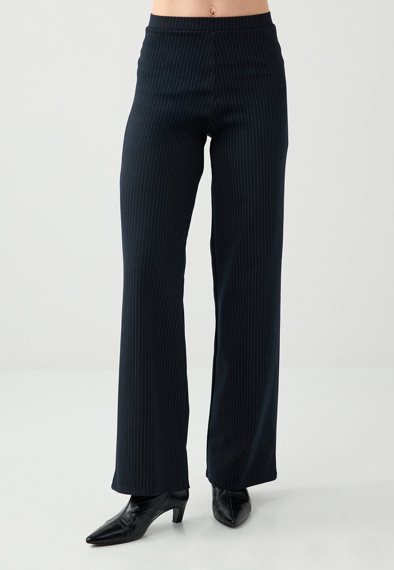 Pantalon noir à fines rayures en tissu lisse, avec une coupe large et une taille haute, associé à des chaussures noires à talons pointues.