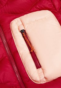 Rode gewatteerde jasbinnenkant met een zachte roze pocket, voorzien van een rits en een oranje trekkoord. "Peak Performance" tekst is in de pocket gedrukt.
