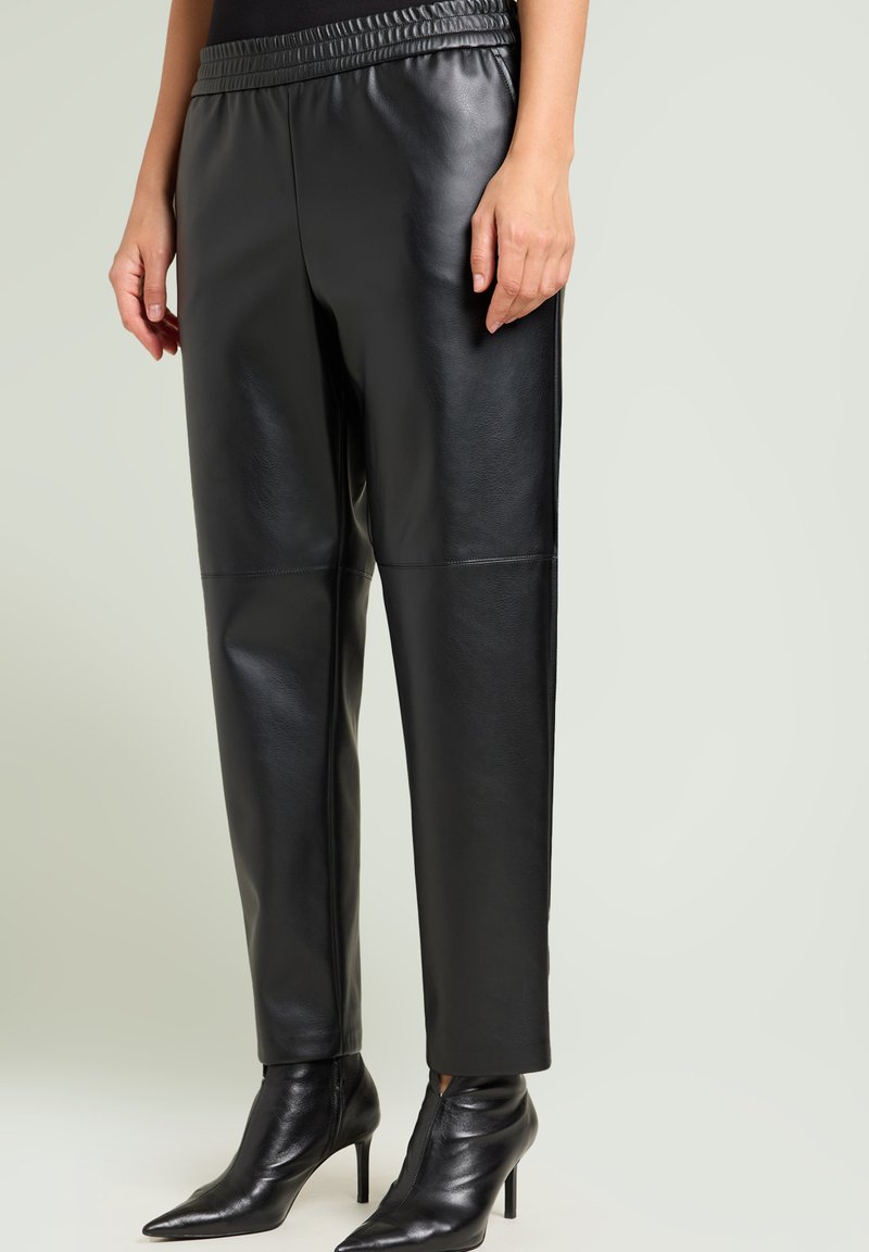 Zwarte imitatieleren broek met een brede elastische tailleband, rechte pasvorm en minimale stikseldetails. Gecombineerd met puntige zwarte enkelboots.