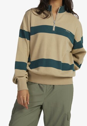 Person iført en beige og mørkegrønstribet quarterzip-trøje og olivengrønne cargobukser, stående med en hånd afslappet