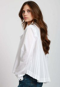 Blusa bianca plissettata con una vestibilità rilassata, dotata di una chiusura frontale con bottoni, maniche lunghe e un design posteriore testurizzato e fluente.