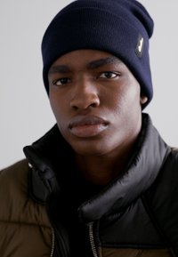 Jūrinė mėlyna megzto beanie skrybėlė ir juodas pūkinis švarkas su aukštu apykakle. Minkšta tekstūra, minimalistinis dizainas, ant skrybėlės yra subtilus logotipo etiketė.