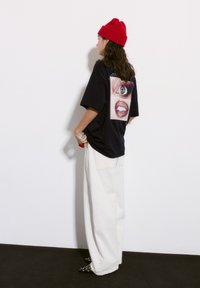 Mustat ylisuuret t-paidat, joissa on selässä silmä- ja huulten graafinen kuvio, yhdistettynä valkoisiin leveälahkeisiin housuihin ja punaiseen pipoon.