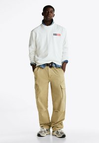 Weißer Sweatshirt mit einem Logo, kombiniert mit einem Jeanshemd. Beigefarbene Cargo-Hosen mit Seitentaschen und graue Sneakers runden das Outfit ab.