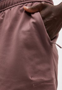 Σκούρο χέρι ακουμπισμένο στην τσέπη μωβ σορτς Nike με ελαστική μέση και ορατό ραμμένο λογότυπο.