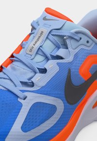 Γαλάζιο και πορτοκαλί παπούτσι τρεξίματος Nike με γκρι λογότυπο swoosh και ετικέτα «Structure Plus» στη γλώσσα, δεμένο με μπλε κορδόνια.