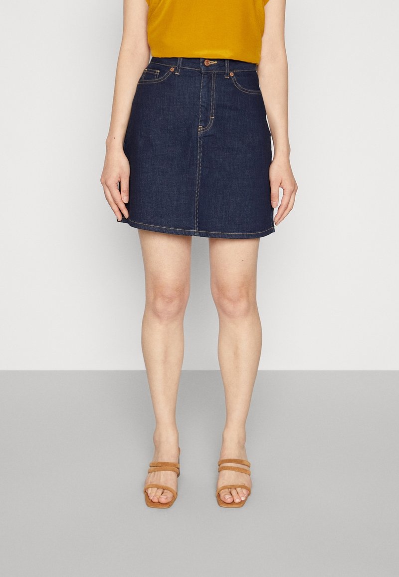 Jupe en denim bleu foncé à coupe droite, avec poches avant et surpiqûres décoratives. Assortie à un haut jaune moutarde et des sandales à talons carrés couleur marron clair.