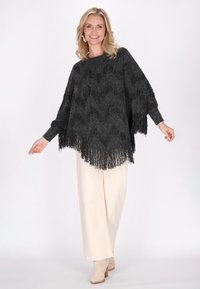 Poncho negru cu franjuri, dintr-un material texturat, având un model în valuri și mâneci largi. Purtați deasupra unor pantaloni largi crem și botine.