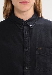 Camicia nera con bottoni, realizzata in tessuto morbido, con colletto, taschino sul petto con etichetta e bottoni a contrasto.