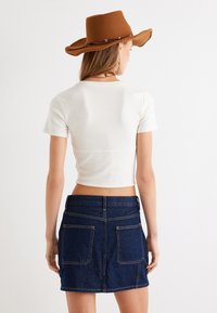 Camiseta blanca corta de mangas cortas combinada con una falda de mezclilla oscura que cuenta con bolsillos traseros y costuras en contraste, llevada con un sombrero marrón.