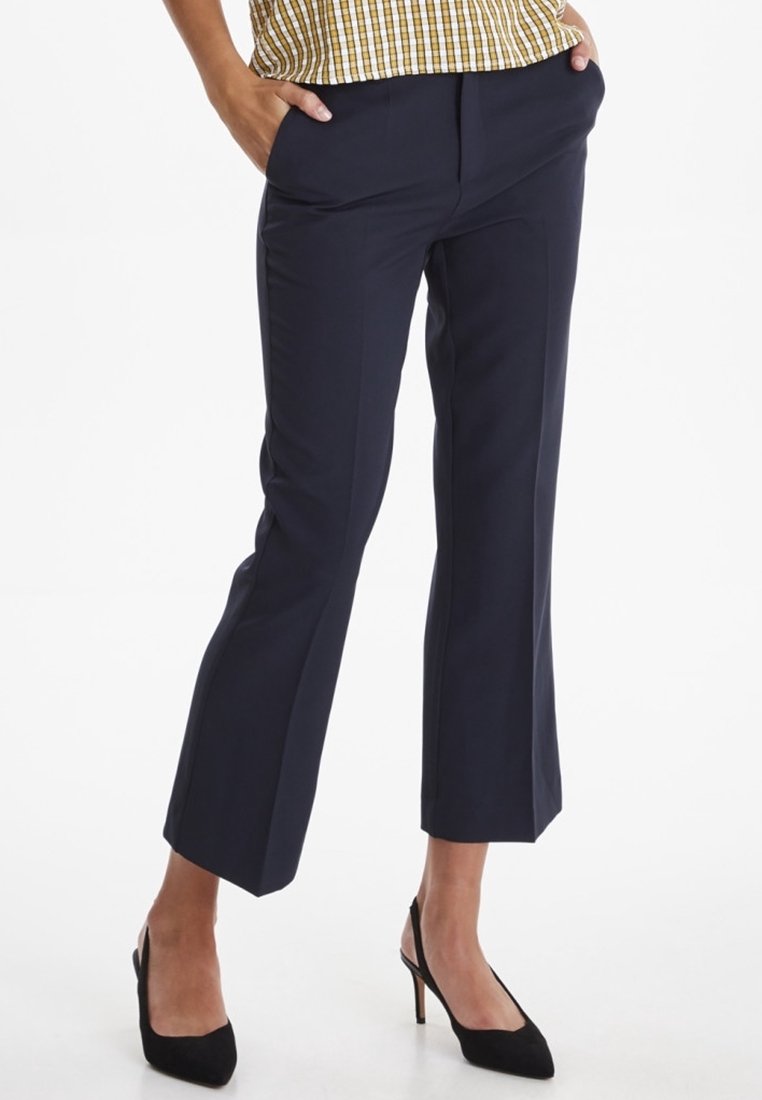 Pantalons ajustés bleu marine à coupe droite, avec des poches latérales et une texture lisse. Portés avec des chaussures à talons noirs.