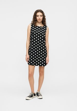 Jeune femme aux cheveux bouclés portant une robe noire sans manches à pois blancs et des baskets noires basses, debout devant un fond uni.