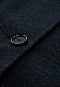 Donker wollen stof met een visgraatpatroon, met een close-up weergave van een zwarte knop, die vier gaten en een gladde oppervlakte toont.