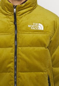 Måste gul senapsgul manchesterpufferjacka med dragkedja, svarta detaljer och broderad logotyp. Har en hög krage och två sidofickor.