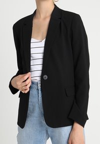 Blazer noir avec revers crantés, fermeture à un bouton et deux poches avant, porté sur un haut rayé blanc et noir.