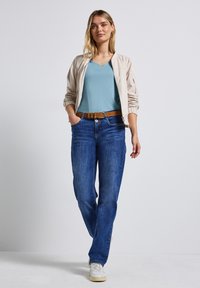 Beige bomberjacka, turkos v-ringad topp, blå denimjeans, brun bälte med silverspänne och vita sneakers. Modell som står med händerna i fickorna.