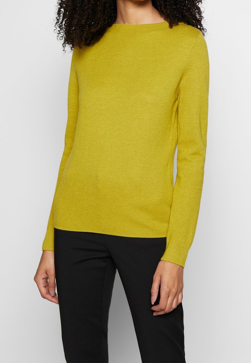 Femme portant un pull en maille jaune moutarde à manches longues et un pantalon noir, debout avec les mains détendues le long du corps.