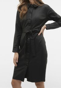 Czarna sukienka z guzikami, wykonana z lekkiego materiału, z kołnierzykiem, długimi rękawami i paskiem w talii, o prostym kroju.