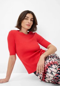 Rode geribbelde top met korte mouwen en een geschulpte halslijn, gecombineerd met een pantalon in patronen van roze, rood en zwarte geometrische vormen.