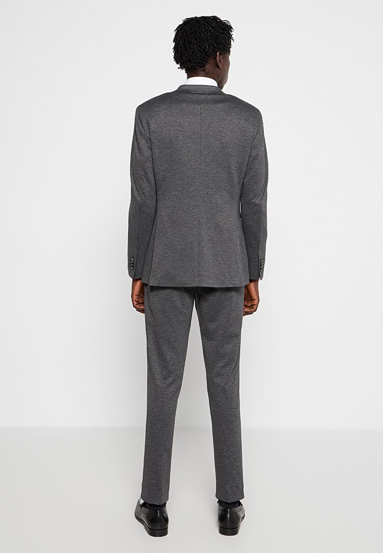 Traje gris con una chaqueta a medida y pantalones a juego. La tela parece suave, con una textura sutil. Zapatos negros completan el atuendo.