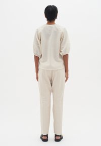 Blusa leggera color crema con maniche a sbuffo, abbinata a pantaloni beige chiaro su misura e sandali neri, vista da dietro.