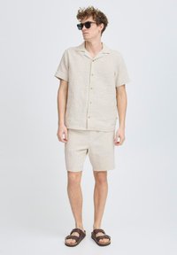 Camisa de manga corta beige y pantalones cortos a juego en tejido texturizado. Cuenta con cierre de botones y sandalias casuales. Diseño simple y ligero.