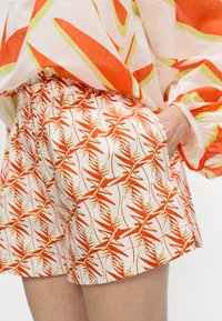 Mønstrede shorts med et livligt orange og gult bladdesign på en hvid baggrund, med elastisk talje og sidelommer.