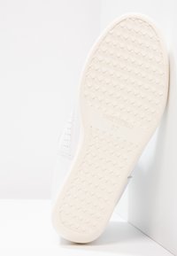 Baskets blanches avec une semelle extérieure en caoutchouc texturée, présentant des détails perforés et un dessus en matériau lisse. L'étiquette de taille indique "37."