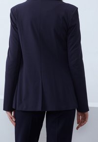 Blazer bleu marine avec une coupe ajustée, une texture lisse et des épaules structurées. Le dos présente une fente unique sans aucune pièce métallique visible ni motifs.