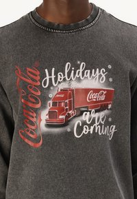 Hanorac gri cu un grafic al unui camion Coca-Cola roșu, cu textul "Sărbătorile vin." Textura moale, decolteu rotund și detalii imprimate.