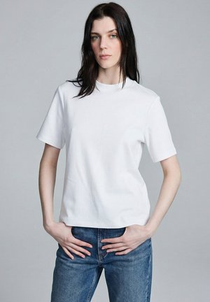 Mujer con cabello largo y oscuro, vistiendo una camiseta blanca lisa de manga corta y vaqueros azules, de pie con las manos cerca de las caderas frente a un fondo gris.