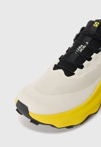 Basket de sport présentant un design crème et jaune, avec un dessus texturé, des lacets noirs et un talon renforcé. Logo proéminent sur le côté.
