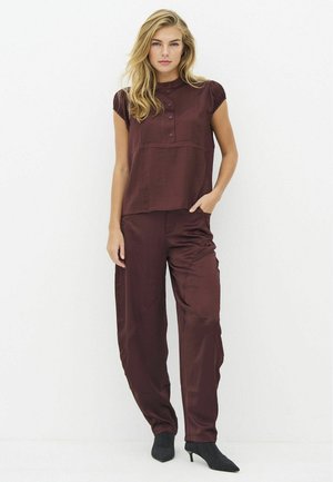 Femme debout sur fond blanc portant un top bordeaux à manches courtes avec boutons et un pantalon large assorti, chaussée de talons noirs pointus.