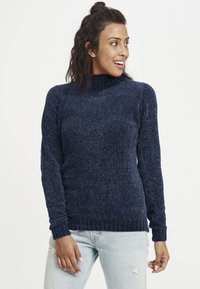 Navyblå strikkegenser med høy ribbestrikket hals, lange ermer og en teksturert, myk overflate. Har en liten logo aksent ved kanten.