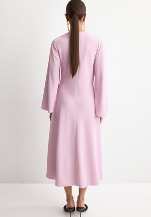 Femme avec une longue queue de cheval brune portant une robe midi à manches longues rose pâle et des mules noires à talons, faisant face à un mur blanc.