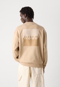 Beige sweatshirt featuring a large square design with the word "HUGO" in varying shades of beige and cream on the back. Soft texture.