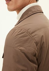 Giacca trapuntata marrone con colletto strutturato, caratterizzata da una leggera lucentezza e dettagli cuciti, indossata sopra un maglione bianco a coste.
