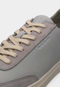 Gros plan d'une basket grise avec des lacets beige et des détails en suède montrant des bords cousus et un petit texte embossé sur le côté.
