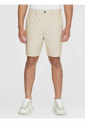 Mand iført beige shorts til knæet med knap og lynlås, parret med hvide sportssko, stående mod en ensfarvet baggrund.