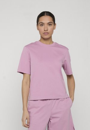 Kobieta z ciemnymi włosami związanymi w kok, ubrana w gładką jasnofioletową koszulkę z krótkim rękawem i dopasowane jasnofioletowe spodnie, z jedną ręką w kieszeni.