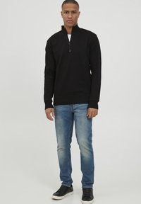 Mustan, pika käistega pullover, millel on pooltagasi tõmmatav krae, kombineeritud siniste, kergelt kulunud teksade ja mustade tossudega.