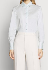 Camicia in satin azzurro chiaro a maniche lunghe con colletto a punta, bottoni verdi e polsini a contrasto, abbinata a pantaloni crema a vita alta.
