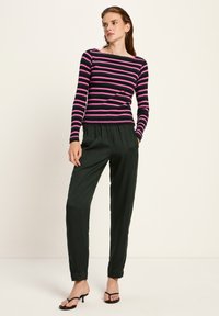 Eine Frau steht da und trägt ein langärmliges Oberteil mit schwarz-pinken Streifen, dunkelgrüne hoch taillierte Hose und schwarze Sandalen mit Absatz, die Hände in den Taschen.