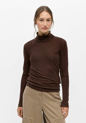 Femme aux cheveux attachés, portant un pull à col roulé marron foncé et un pantalon beige, debout devant un fond blanc uni.