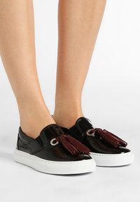 Chaussures à enfiler en cuir verni noir avec accents de pompons rouges, semelle en caoutchouc blanc et œillets métalliques ronds pour les lacets. Design plat pour un usage décontracté.