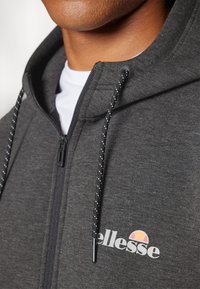 Šedá mikina na zips s čierno-bielymi šnúrkami, obsahujúca grafické logo. Tkanina pôsobí mäkko s hladkou textúrou.