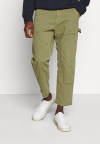 Man in olijfgroene cropped cargo broek en witte sneakers, staand met één hand in zijn zak tegen een effen achtergrond.