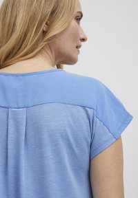Krátký rukáv top v světle modré tkanině s hladkou texturou. Disponuje kontrastním designem na ramenou a jemnými sklady na zadní straně.