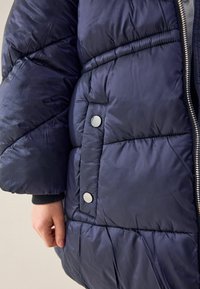 Tengerész kék puffertakaró, steppelt dizájnnal, fényes textúrával, ezüst színű nyomógombokkal és látható cipzárral. Puha, szigetelt anyag.