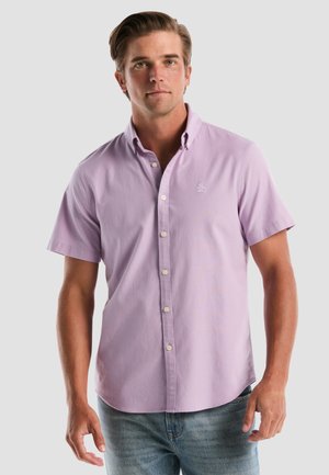 Man draagt een lila kortermig overhemd met knopen en een klein geborduurd logo op de borst, gecombineerd met lichtblauwe jeans.